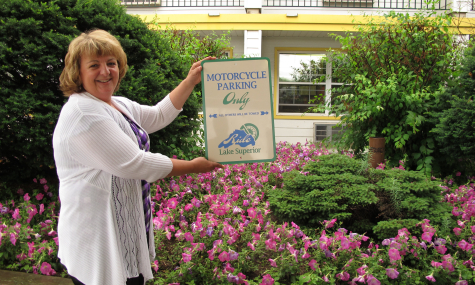 woman wearing a white sweater, holding a sign standing in front of flower garden with pink petunias